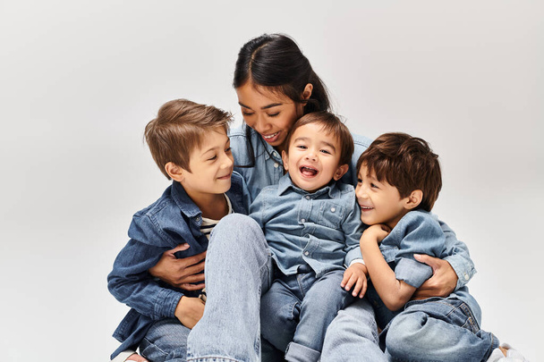 Een groep kinderen, waaronder een jonge Aziatische moeder en haar zoons, in denim kleding, zitten op elkaar in een grijze studio. - Foto, afbeelding