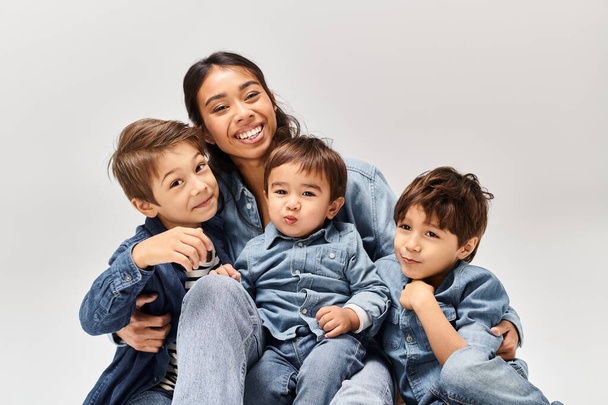 Una giovane madre asiatica vestita di denim siede con i bambini, i suoi piccoli figli anche in denim, in uno studio grigio. - Foto, immagini