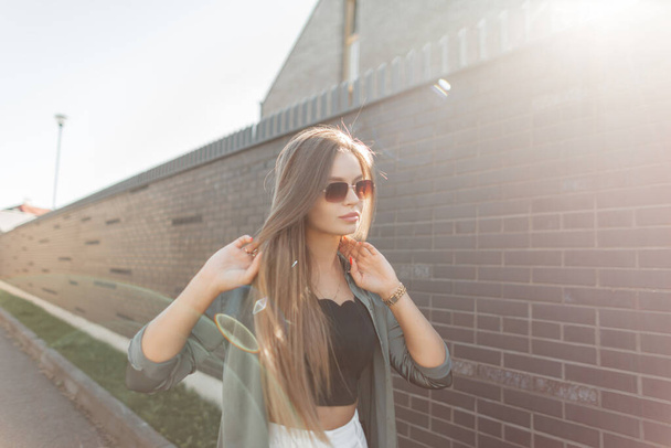 Belle jeune fille d'affaires réussie avec des lunettes de soleil en vêtements à la mode se promène près d'une clôture en brique noire au coucher du soleil. Jolie femme - Photo, image