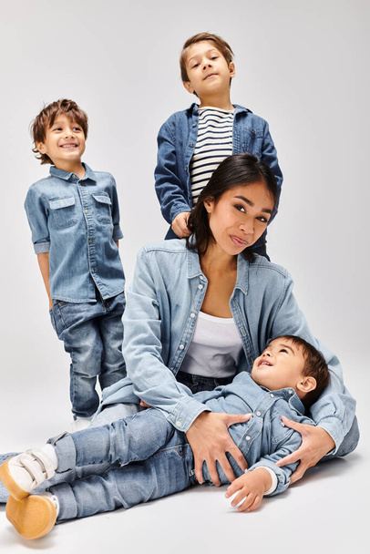 Genç bir Asyalı anne, gri bir stüdyoda, kot giyip oğullarıyla birlikte yerde oturuyor.. - Fotoğraf, Görsel