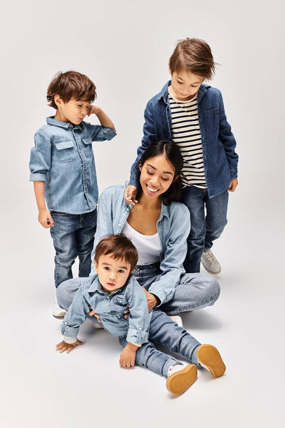 Una joven madre asiática y sus hijos pequeños, todos vestidos de mezclilla, posando para una foto en un estudio gris. - Foto, Imagen