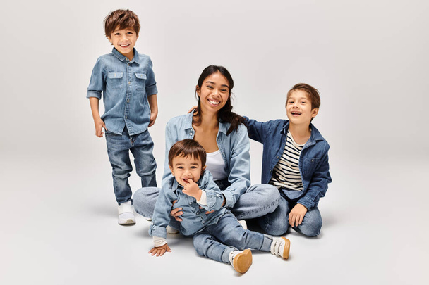 Una giovane madre asiatica e i suoi piccoli figli indossano abiti in denim in posa insieme in un elegante studio grigio. - Foto, immagini