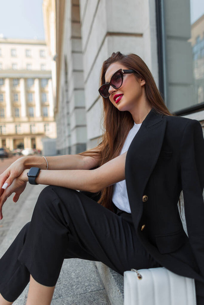 Menina de negócios feliz urbana bonita elegante com óculos de sol de moda na moda roupas casuais pretas elegantes com blazer sentado em passos na cidade - Foto, Imagem