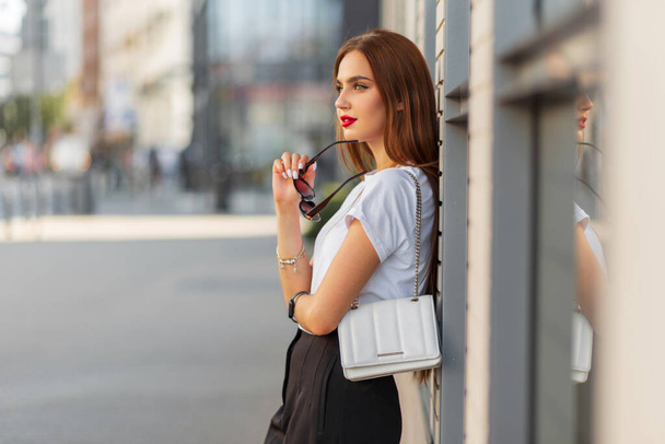 Κομψό όμορφο κορίτσι της αστικής μόδας σε ένα μπλουζάκι μόδας με μια μοντέρνα λευκή τσάντα με γυαλιά ηλίου κοντά σε έναν τοίχο της πόλης. Όμορφη κυρία. - Φωτογραφία, εικόνα