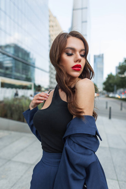 Glamoureuze mooie mode meisje met rode lippen in een stijlvol blauw pak met een zwarte top wandelingen in de moderne stad in de buurt van het kantoorgebouw - Foto, afbeelding
