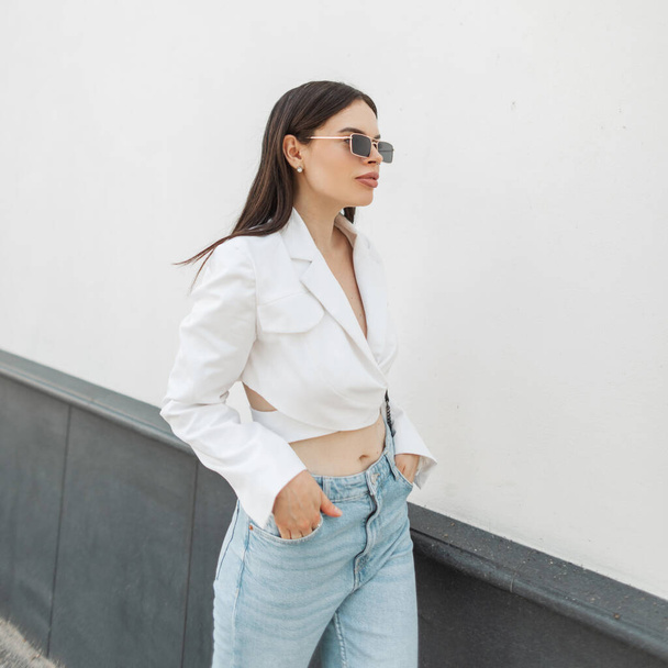 Стильная красивая модная женщина модель в старинных солнцезащитных очках в модном белом пиджаке в синих джинсах гуляет по улице возле белой стены - Фото, изображение