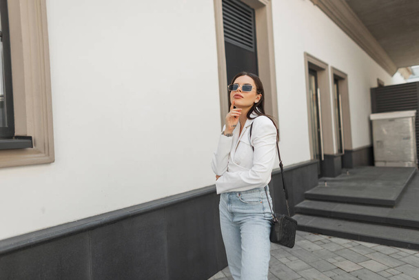 Divatos városi divat szépség nő modell napszemüveg fehér elegáns blézer és divatos kék farmer táska séták a városban - Fotó, kép