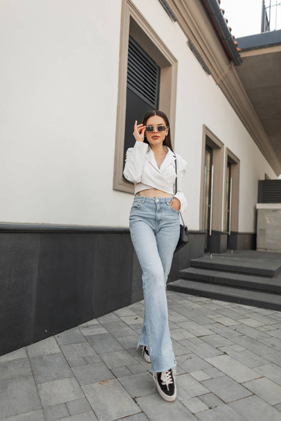 Cool bella ragazza caucasica alla moda in abiti casual urbani di moda con una giacca bianca alla moda, jeans e scarpe da ginnastica indossa occhiali da sole vintage e passeggiate in città - Foto, immagini