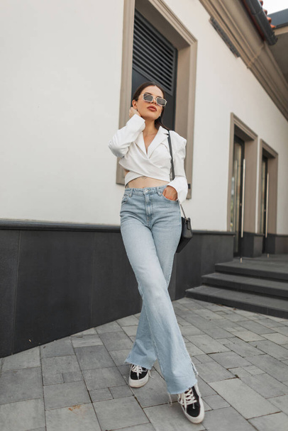Modelo de mulher urbana bonita elegante com óculos de sol em uma roupa casual de moda com uma jaqueta, jeans vintage azul e tênis com uma bolsa fica na rua - Foto, Imagem