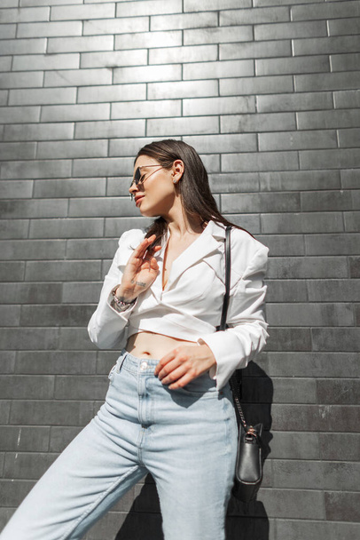 Elegante bella ragazza caucasica urbana con occhiali da sole vintage in abiti casual di moda con una giacca bianca, jeans blu con una borsa vicino a un muro di mattoni alla luce del sole - Foto, immagini