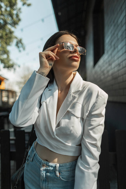 Piękna kobieta mody w modnych ubraniach casual z białą kurtką stawia na zabytkowe okulary przeciwsłoneczne i spacery w mieście w słońcu - Zdjęcie, obraz