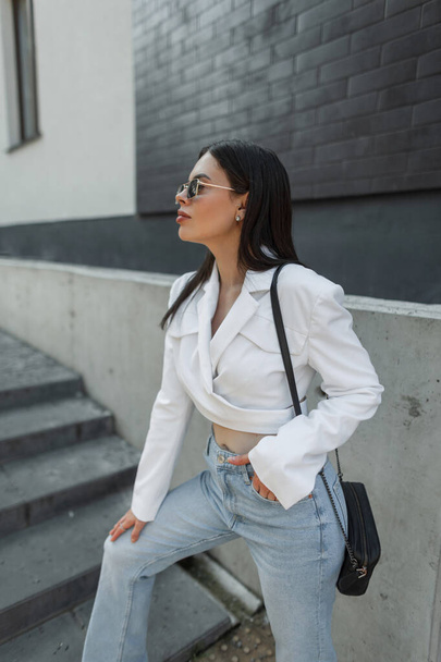 Menina elegante bonita na moda com óculos de sol na moda roupas casuais com um blazer branco e jeans azul vintage com uma bolsa na cidade perto de uma parede preta de tijolo - Foto, Imagem