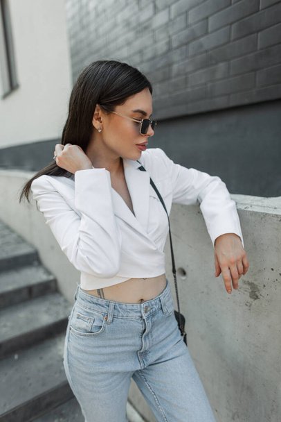 Bella elegante modello glamour ragazza di moda con occhiali da sole alla moda in un blazer bianco alla moda e jeans blu si trova vicino a un edificio di mattoni neri in città - Foto, immagini