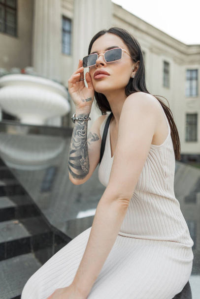 Beautiful stylish hipster girl with vintage sunglasses in a fashion beige dress with a tattoo on her arm sits in the city near the columns building. Trendy pretty lady - Photo, Image