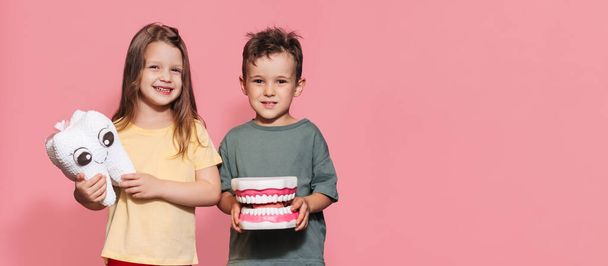 Een lachende jongen en een meisje met gezonde tanden houden een speelgoedtand in hun handen op een geïsoleerde achtergrond. Een plek voor je sms. Orale hygiëne. Pediatrische tandheelkunde. Protheses. Regels voor het poetsen van tanden - Foto, afbeelding