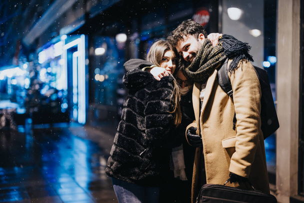 Ein liebevolles junges Paar, das sich auf einer schneebedeckten, beleuchteten Stadtstraße zusammenkuschelt, schafft trotz des kühlen Wetters eine Atmosphäre der Romantik und Wärme. - Foto, Bild
