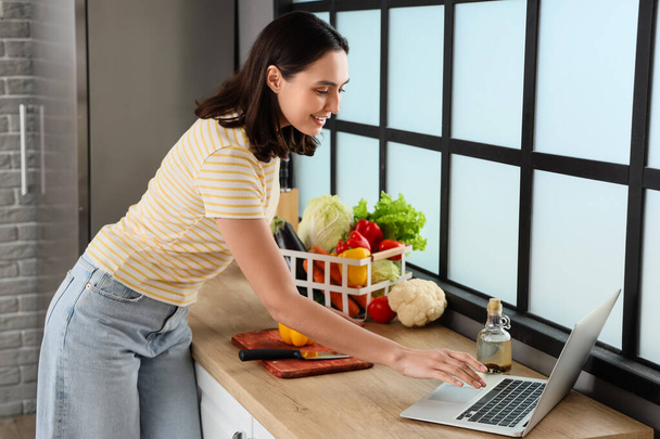 Jonge vrouw met verse groenten met laptop in de keuken - Foto, afbeelding