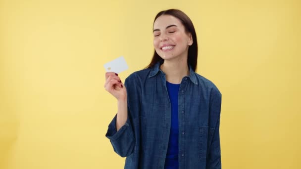 Portrait of smiling caucasian woman pointing with index finger on white bank card holding in hand over yellow background. Female brunette recommending cashless payment for convenient shopping. - Footage, Video