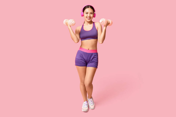 Jonge vrouw in sportkleding luisteren naar muziek en training met halters op roze achtergrond - Foto, afbeelding