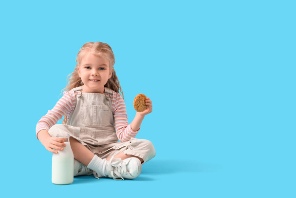 青い背景に座っている牛乳とクッキーのボトルを持つかわいい少女 - 写真・画像