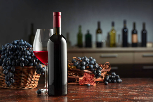 Bottle and glass of red wine with blue grapes on a vintage table. In the background bar counter with various wine bottles. Focus on a foreground. - Photo, Image