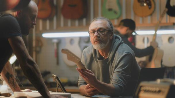 Yetişkin bir erkek, gitar planları çizmek için dijital tablet bilgisayar kullanır. Orta yaşlı dövmeli marangoz iş arkadaşıyla konuşuyor, gitar fretboardları alıyor. Zanaatkarlar şık bir atölyede çalışırlar.. - Fotoğraf, Görsel