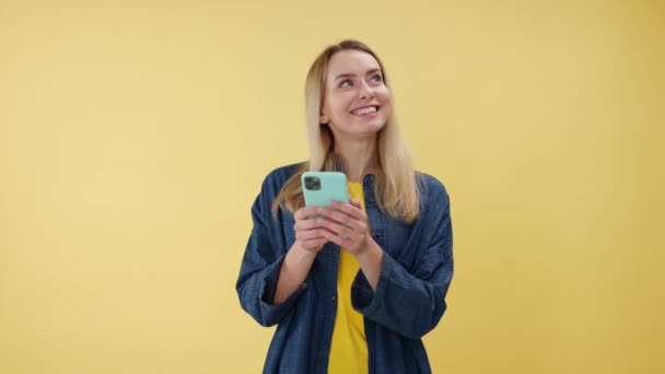 Yüzünde gülümseme olan güzel beyaz bir kadın kapalı mekanda sohbet etmek için akıllı telefon kullanıyor. İnternette mesaj yazan ve sosyal medyada gezinen hoş bir kadın. Sarı arkaplan üzerinde izole edilmiş. - Video, Çekim