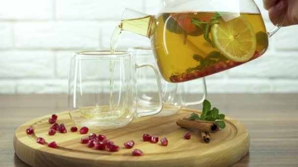Una mano sosteniendo una tetera de vidrio transparente vierte té de frutas calientes en una taza de vidrio colocada sobre la mesa, té de invierno con limón y naranja, palitos de canela, especias y bayas, cámara lenta - Imágenes, Vídeo
