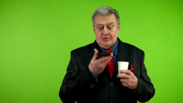 Senior man in suit drinking coffee and records voice message with smartphone. Using smart technologies for social media communication, voice commands, messaging. Green screen, chroma key. - Footage, Video