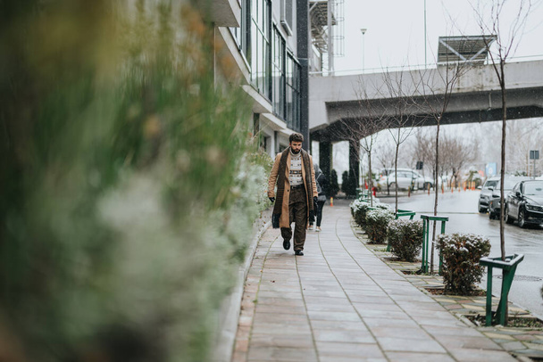 Een eenzame jonge volwassene trotseert een besneeuwde dag, wandelt op de stoep van de stad met een focus en vastberadenheid, toont het stedelijke winterleven. - Foto, afbeelding