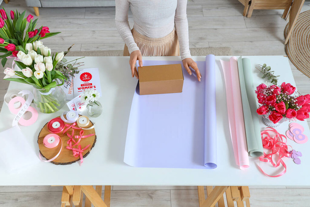Женщина с подарочной коробкой, упаковочные материалы и красивые цветы тюльпан за светлым столом. Международный женский день - Фото, изображение