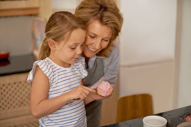 Woman, child and icing cupcake in kitchen for baking decoration with grandmother, bonding or ingredients. Female person, girl and recipe for sweet dessert at counter or teaching, helping or support. - Photo, Image
