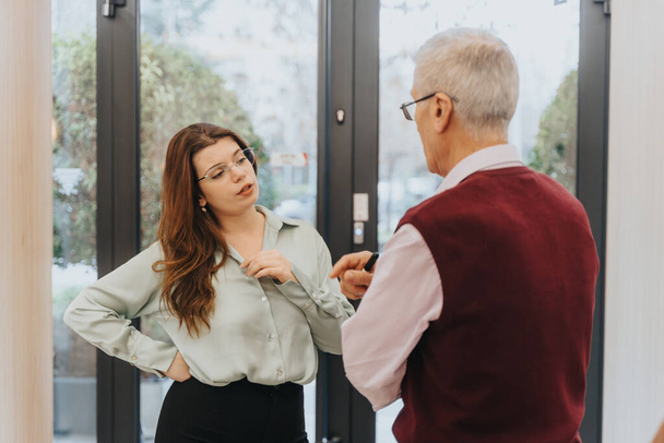Two, different aged people, coworkers, engage in a conversation by the office doorway. The woman, with a questioning stance, discusses with the older man. - Photo, Image