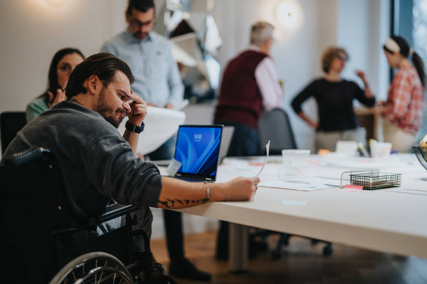 Συγκεντρωμένη ομάδα διαφορετικών επαγγελματιών, συμπεριλαμβανομένου ενός άνδρα σε αναπηρική καρέκλα, που συνεργάζεται σε ένα σύγχρονο περιβάλλον γραφείου. - Φωτογραφία, εικόνα