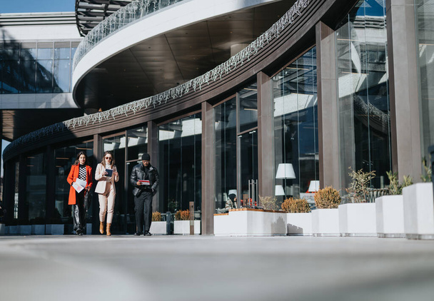 Due professioniste e un dirigente di sesso maschile camminano e conversano sullo sfondo di un edificio per uffici contemporaneo, trasmettendo un senso di vita aziendale e di lavoro di squadra. - Foto, immagini