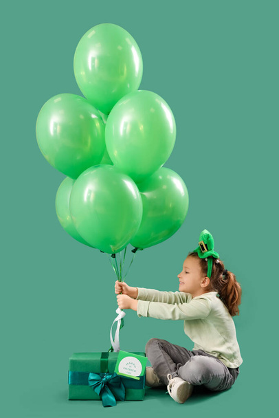 Linda niña con sombrero de duende, caja de regalo y globos sobre fondo verde. Celebración del Día de San Patricio - Foto, imagen
