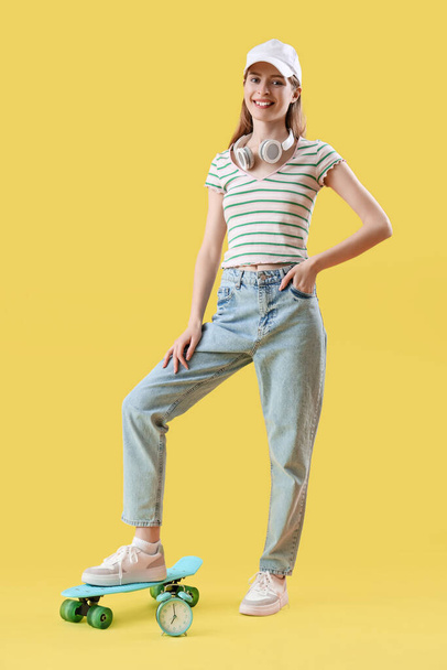 Young woman with skateboard and alarm clock on yellow background - Photo, Image