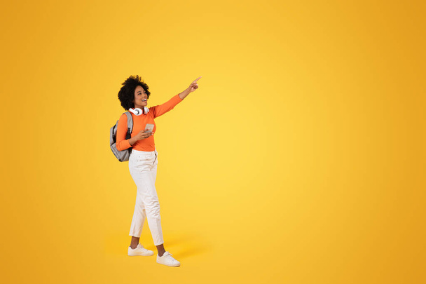 Mujer afroamericana alegre con el pelo rizado, señalando con emoción, sosteniendo un teléfono, usando una camiseta naranja, pantalones blancos, auriculares y una mochila, sobre un fondo amarillo brillante - Foto, imagen