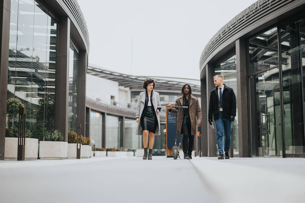 Un grupo diverso de jóvenes colegas camina confiadamente fuera de un moderno edificio de oficinas, demostrando trabajo en equipo y colaboración. - Foto, Imagen