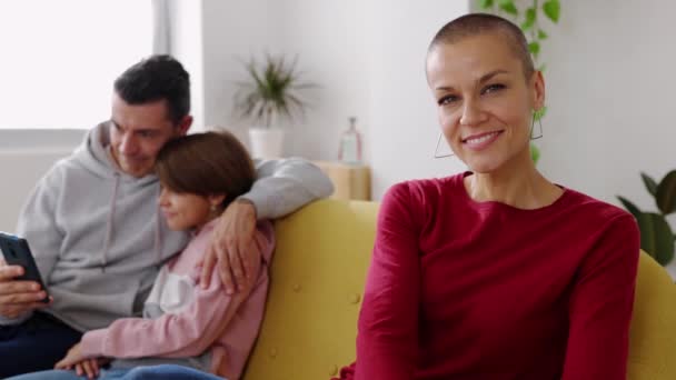 Портрет усміхненої середньої дорослої жінки, яка посміхається на камеру, сидячи на дивані з сім'єю, використовуючи телефон на фоні - Кадри, відео