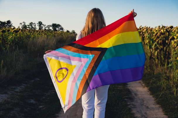 Σύμβολο του μήνα υπερηφάνειας ΛΟΑΤΚΙ. Νεαρή γυναίκα δείχνει Rainbow LGBTQIA σημαία κυματίζει στον άνεμο κατασκευασμένο από μετάξι υλικό σε φόντο τομέα. Ίσα δικαιώματα. Έννοια ειρήνης και ελευθερίας - Φωτογραφία, εικόνα