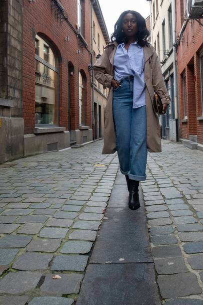 この写真は,若いアフリカ人女性が石畳の路地でカメラに向かって歩き,自信と独立感を呼び起こします. 青いボタンダウンシャツで構成された彼女の服 - 写真・画像