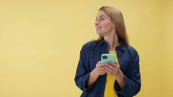 Smiling caucasian woman browsing personal smartphone over yellow studio background. Emotional female looking aside while using modern gadget to keeping up with time indoors. Empty space for text. - Footage, Video