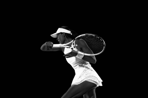 Athletic woman in tennis attire playing backhand against black studio background. Monochrome filter. Concept of women in sport, active lifestyles, tournaments and events, energy, movement. - Photo, Image