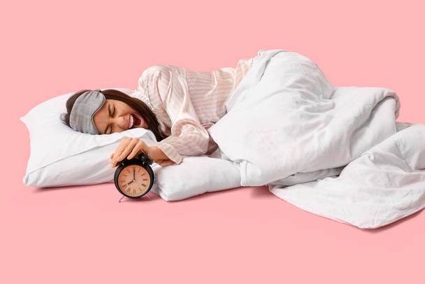 Θυμωμένη νεαρή γυναίκα με κουβέρτα κλείνοντας το ξυπνητήρι σε ροζ φόντο - Φωτογραφία, εικόνα