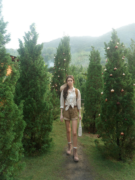 A woman in a white shirt and brown shorts walking through a forest of evergreen trees - Photo, Image
