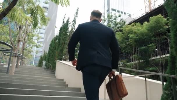 Widok na białego menadżera w garniturze wchodzącego po schodach, trzymającego walizkę w zielonym mieście. Profesjonalny biznesmen idzie do pracy podczas wspinaczki po schodach w eko-miejskim mieście. Urban.. - Materiał filmowy, wideo