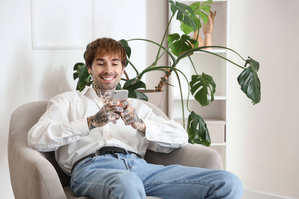 Jóképű férfi mobiltelefont használ puha karosszékben otthon - Fotó, kép
