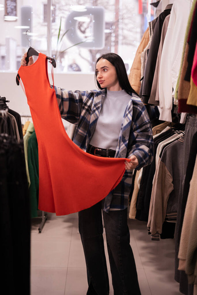 A fiatal nő ruhát választ a plázában vagy a ruhaboltban, és narancssárga ruhát keres. vásárlás, divat, stílus és emberek koncepció. Fénymásolási hely - Fotó, kép
