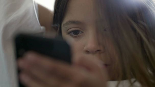 Jedna dziewczynka korzystająca z telefonu komórkowego wpatrująca się w ekran, z bliska obcisła twarz dziecka zaangażowanego w nowoczesne technologie przewijania mediów online - Zdjęcie, obraz